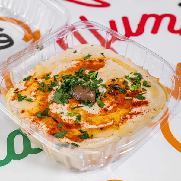 Yalla Shawarma's hummus in a clear plastic container, garnished with paprika, parsley, and an olive.