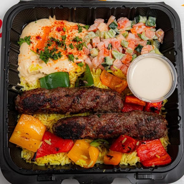 Two grilled kofta kebabs on yellow rice with hummus, salad, bell peppers, and a side of sauce.